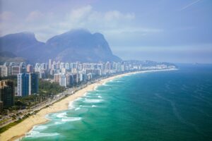 Vista aérea da praia e skyline da Barra da Tijuca no Rio de Janeiro, com prédios residenciais e a Pedra da Gávea ao fundo