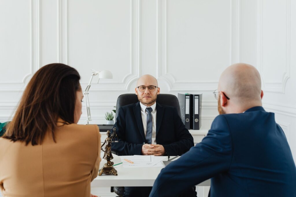 Advogado em consulta com casal de clientes em escritório jurídico, com estátua da Justiça sobre a mesa, representando o atendimento em planejamento sucessório.